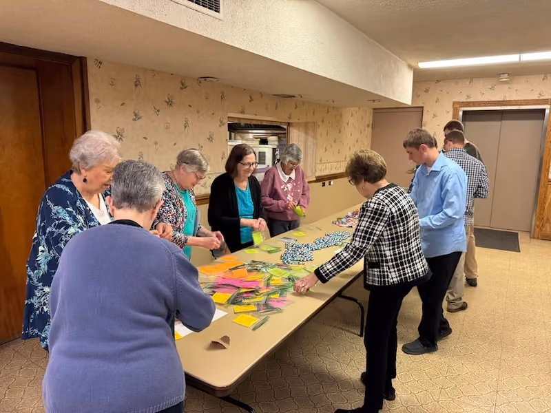 A group of elderly and middle-aged people standing around a long table indoors, sorting or organizing colorful paper packets and items spread out on the table in a carpeted room with beige floral wallpaper.