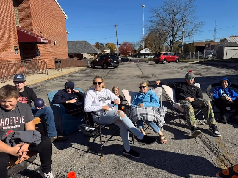 Seven people sitting outdoors on chairs in a parking lot near a red brick building with a maroon canopy labeled Entrance, under clear blue sky.