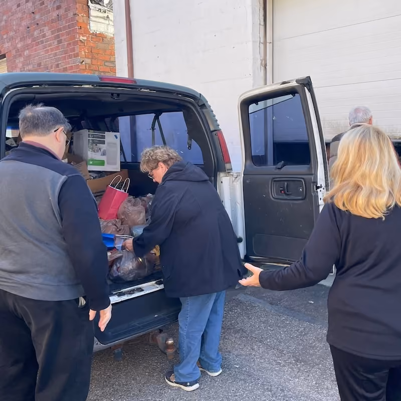 Four adults unloading bags of groceries and other items from the back of a white van parked near a brick and white wall loading area.
