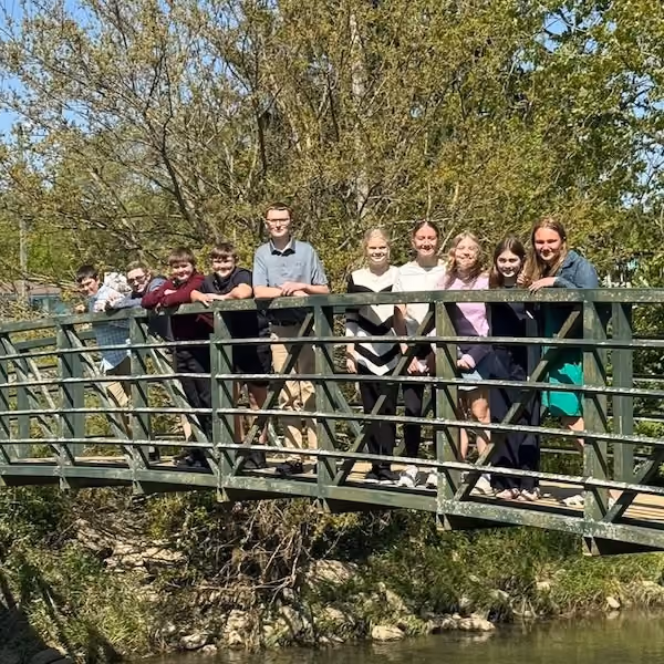 A group of ten young people standing together on a green metal footbridge over a shallow river, surrounded by trees with green leaves on a sunny day.