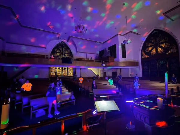 Interior of a church with wooden pews and stained glass windows, illuminated by colorful disco lights on the ceiling and walls, with a group of young people, some in bright clothing, seated and standing spaced apart.