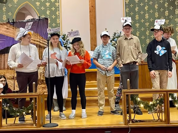 Group of six youth standing on a church stage, each holding papers and wearing hats with signs displaying numbers and words, in front of a decorated altar with flowers and garlands.