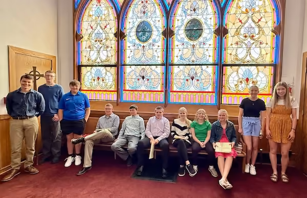 Group of eleven smiling youth, six sitting on a wooden bench and five standing, in front of a large colorful stained glass window inside a church with red carpet.