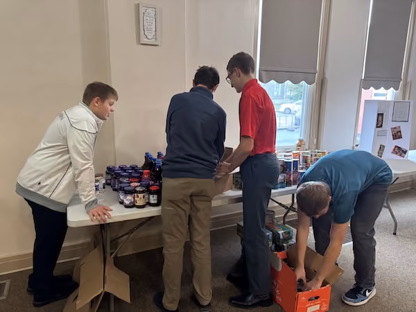 Four young men organizing and packing jars and cans of food on and under a folding table in a room with windows and a bulletin board.