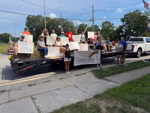 Group of people sitting and standing on a flatbed trailer attached to a white pickup truck, holding handmade signs and a banner reading Good Hope Lutheran Church.