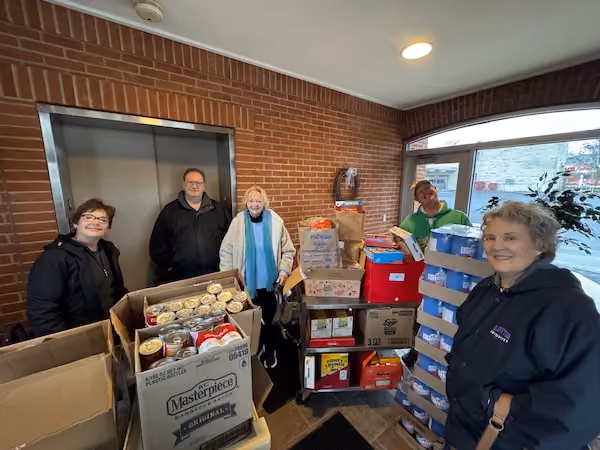 Five people standing inside a brick entryway next to boxes and packs of canned and boxed food items, smiling at the camera.