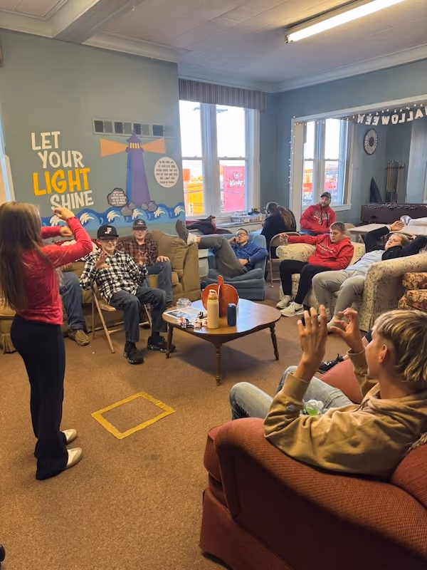 A group of young people gathered in a cozy room with couches and chairs, some sitting and some standing. A girl in a red jacket is standing and gesturing, while others watch and interact. The wall features a mural with a lighthouse and the phrase 'Let Your Light Shine.' The room is well-lit with large windows and decorated with string lights.