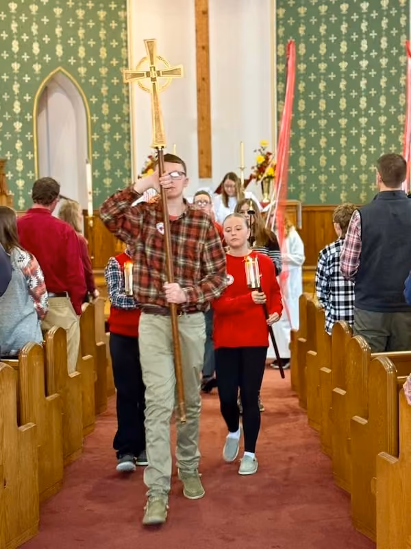 Young people processing down the central aisle of a church holding candles and a tall cross, with wooden pews and green patterned walls in the background.