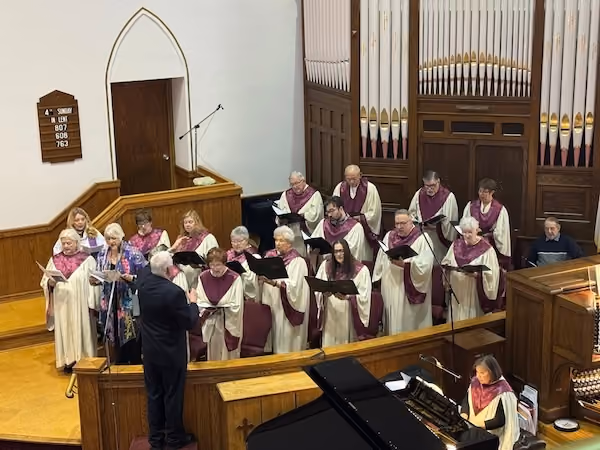 Church choir in cream robes with purple stoles singing in a church sanctuary, led by a conductor in a black suit, with an organist playing at the organ and a black grand piano visible.