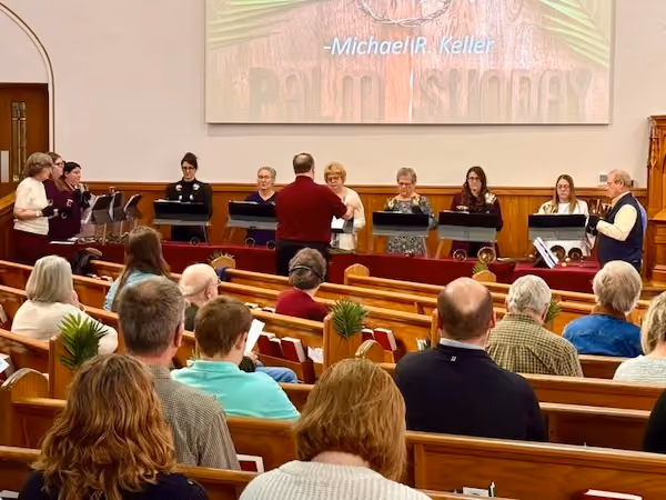 Congregation seated in wooden pews watching a handbell choir perform at the front of a church sanctuary, with a conductor leading them and a projector screen displaying "Palm Sunday" behind.