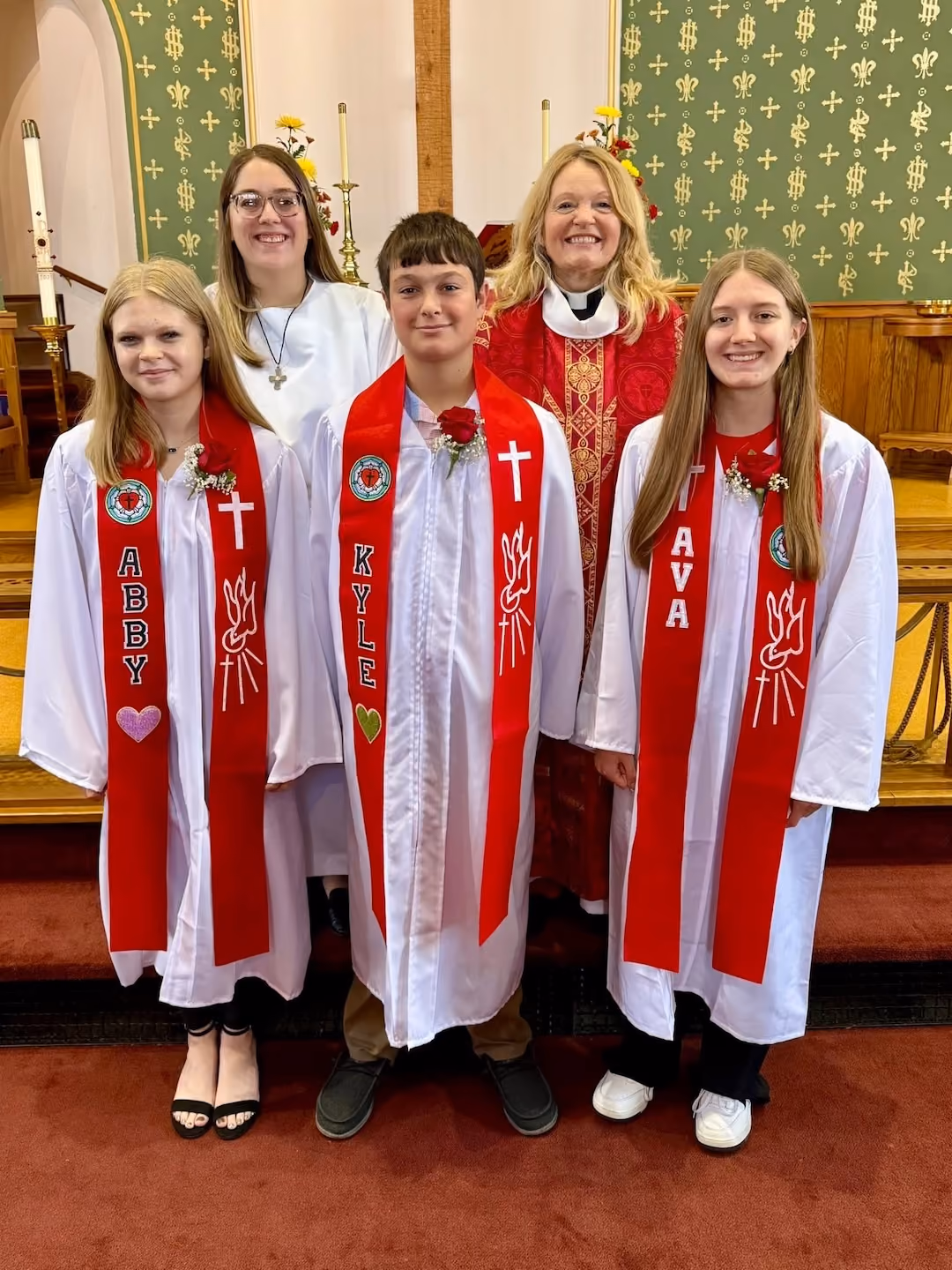 Three young people named Abby, Kyle, and Ava wearing white robes and red stoles with their names stand in front of two women also in religious attire inside a church, with candles and green altar cloths in the background.