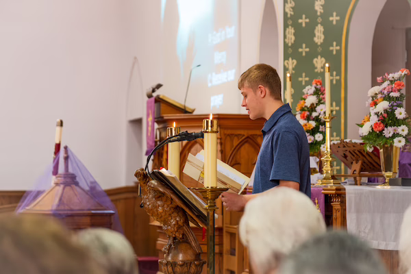 A young man in a blue shirt reading from the Bible at a church pulpit decorated with candles and flowers, with blurred heads of an audience in the foreground.