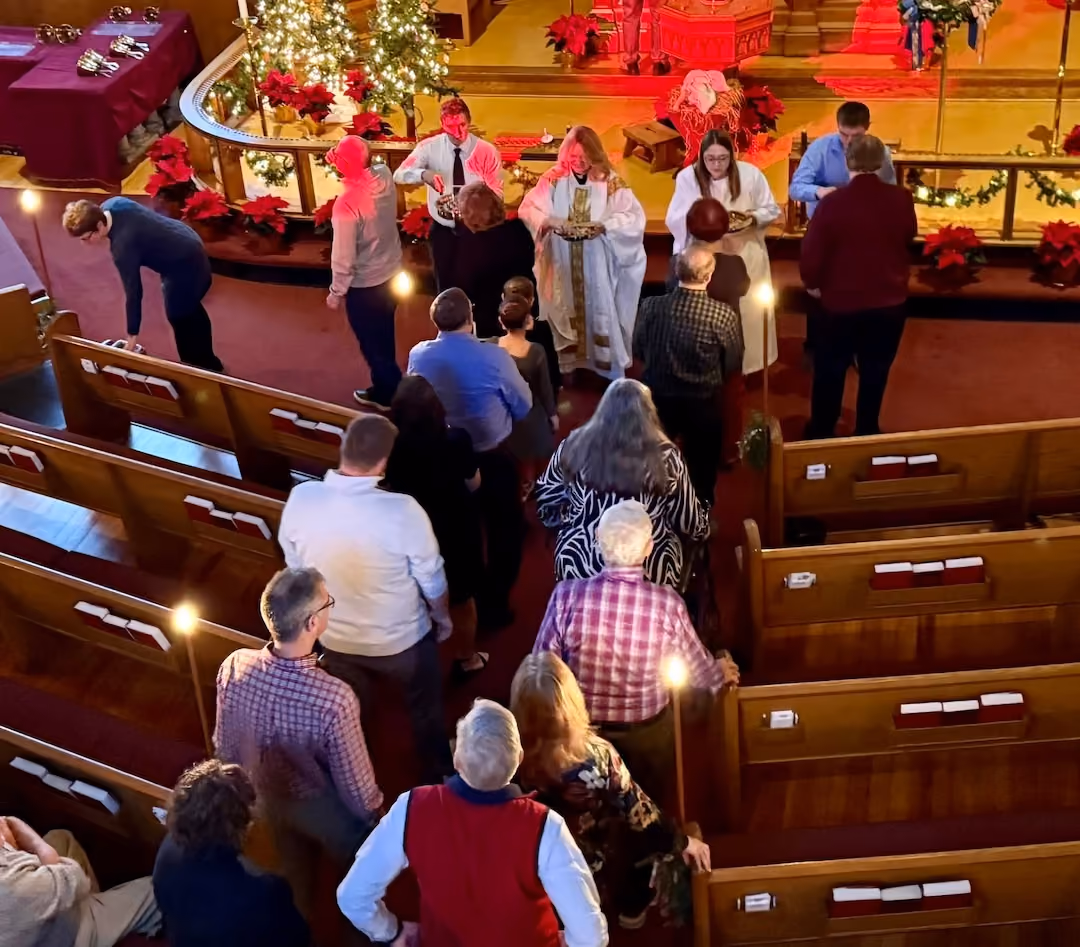 Congregants in a church lined up in aisles receiving communion from two ministers dressed in white robes, with Christmas decorations including poinsettias and lit trees around the altar area.