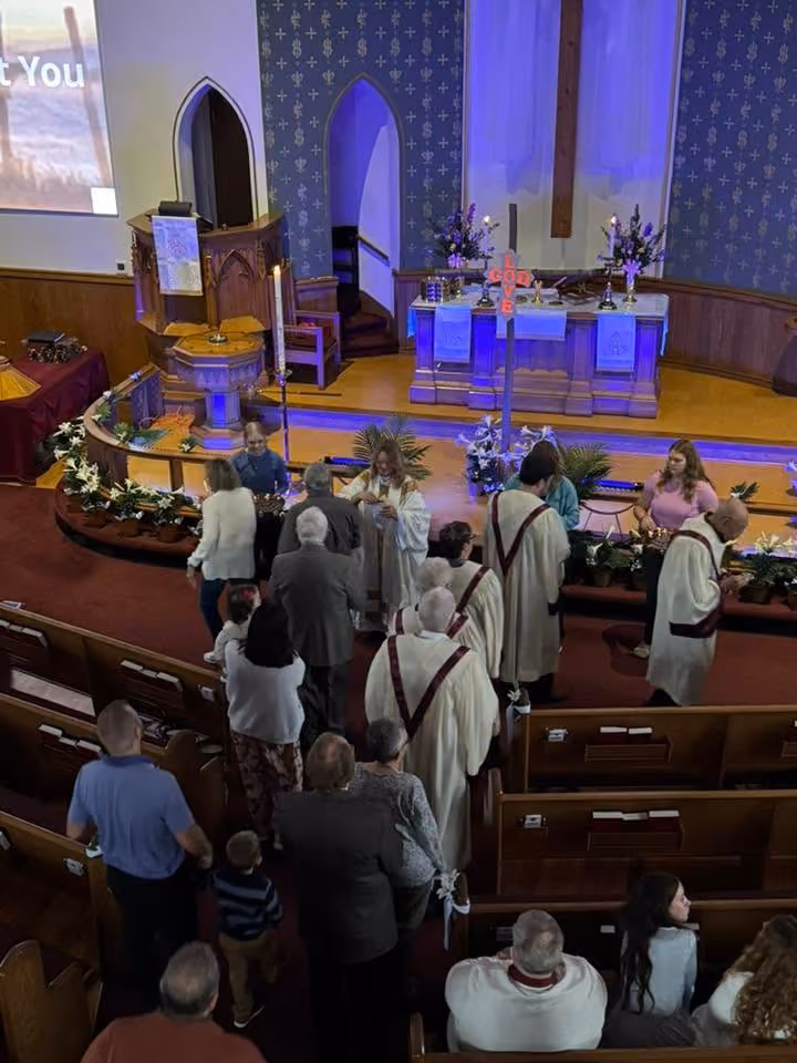 Congregation members receive communion in a church sanctuary near an altar decorated with flowers and a large cross.