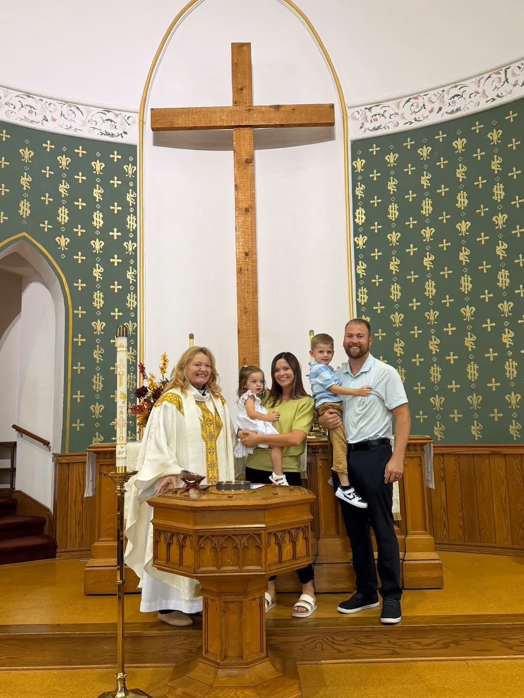 A family of four with a woman clergy in white and gold robes stand in front of a large wooden cross inside a church for baptism; the mother and father each hold a child, with green patterned walls and church furniture in the background.