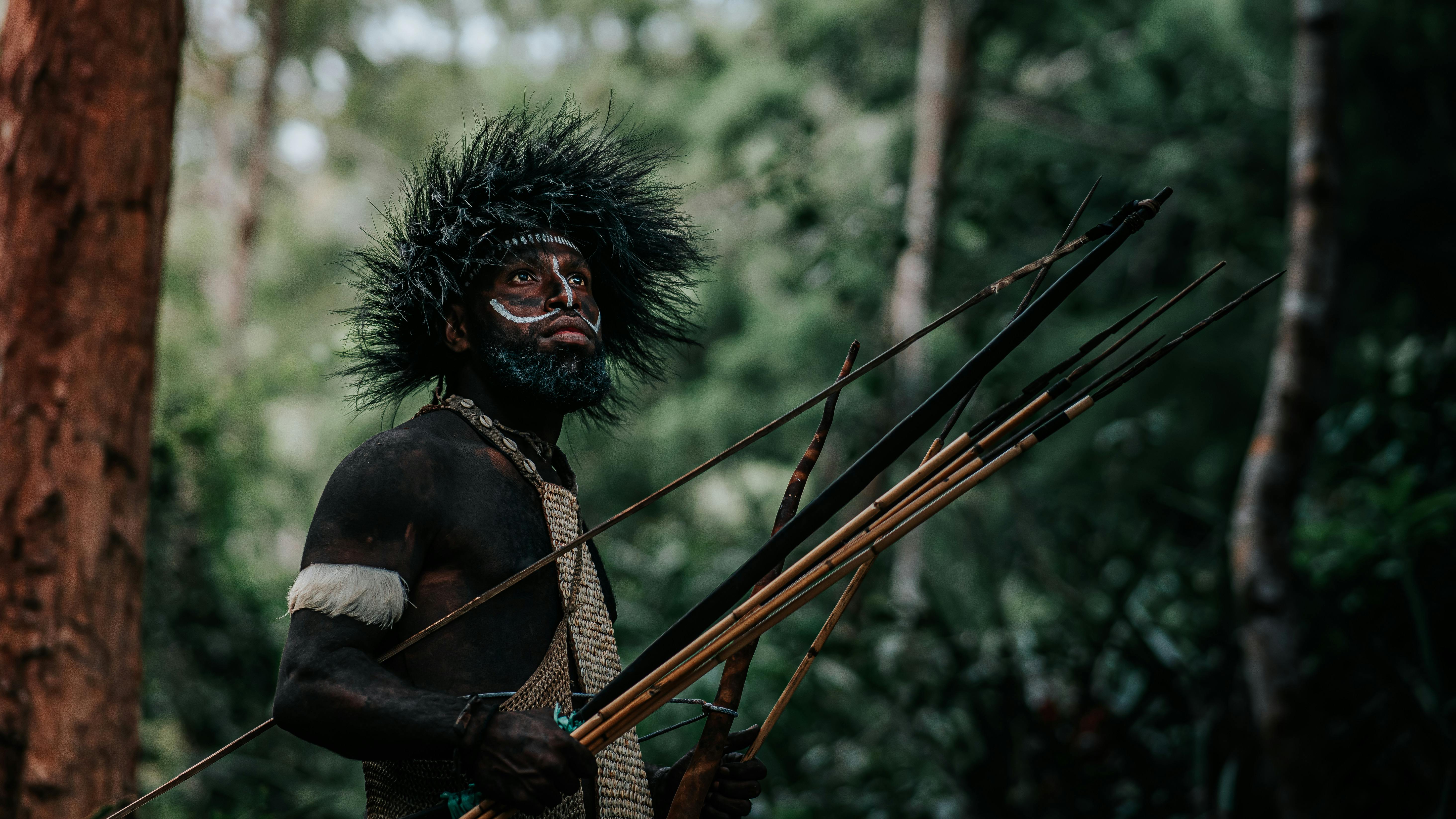 Indigenous man with tribal face paint and feathered headdress holding multiple spears in a forest setting.