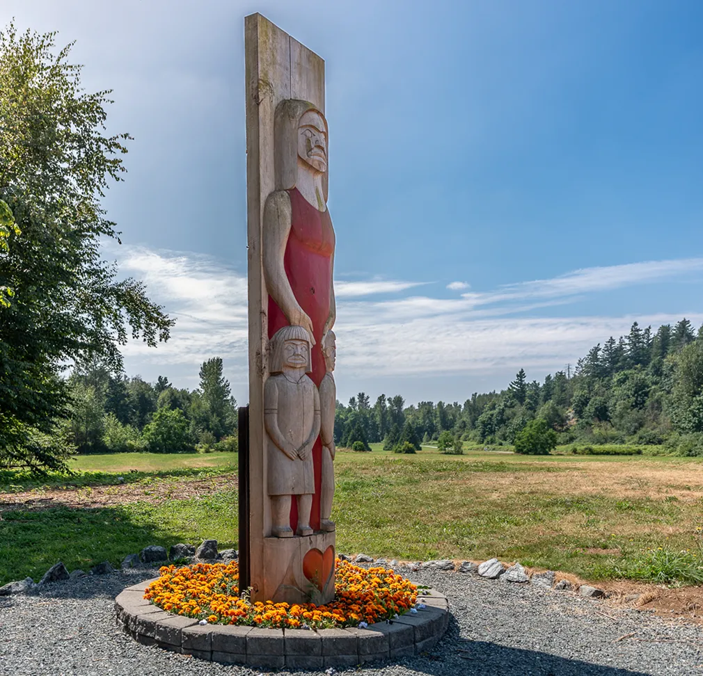 Wooden totem pole carved with stylized human figures, standing on a circular stone base surrounded by orange flowers in a grassy field under a blue sky.