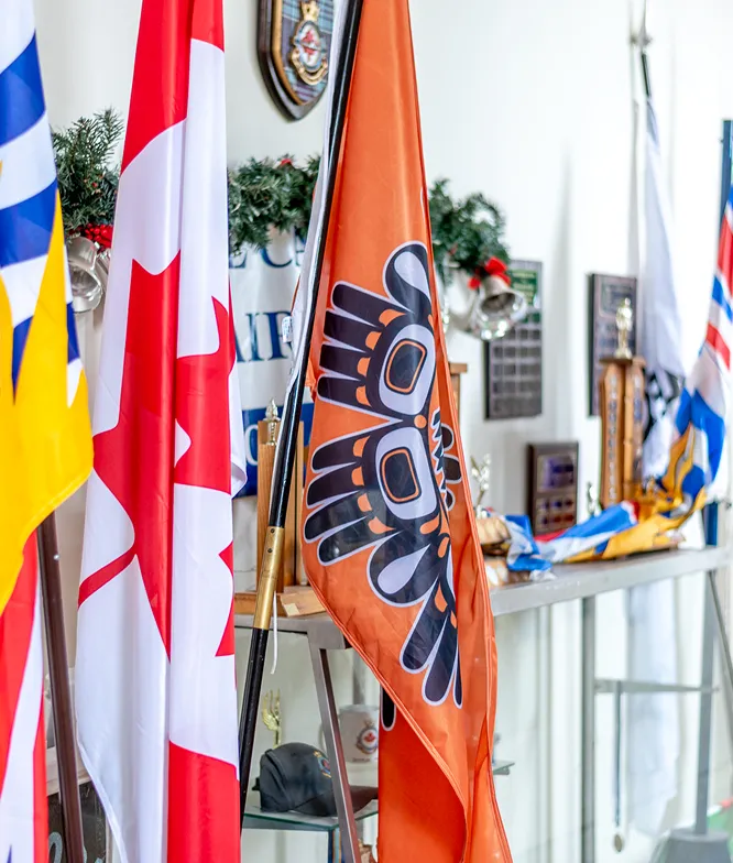 Close-up of various flags including a Canadian flag and an orange flag with Indigenous art, displayed indoors near trophies and plaques.