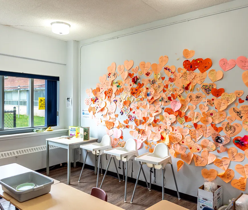 Bright classroom corner with three white high chairs in front of a wall decorated with numerous orange and pink heart-shaped notes and crafts.