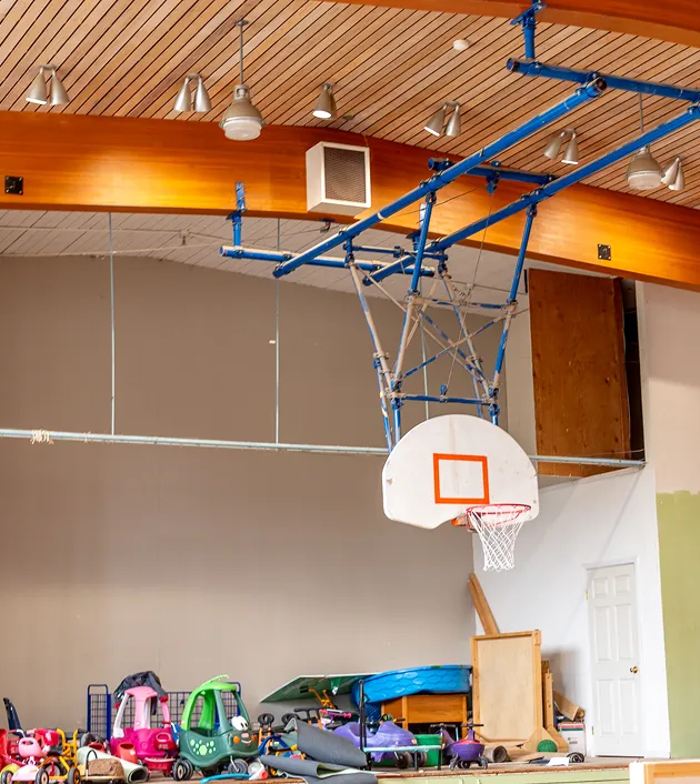Indoor basketball hoop mounted on a blue metal frame with wooden ceiling and a pile of colorful children's ride-on toys beneath it.
