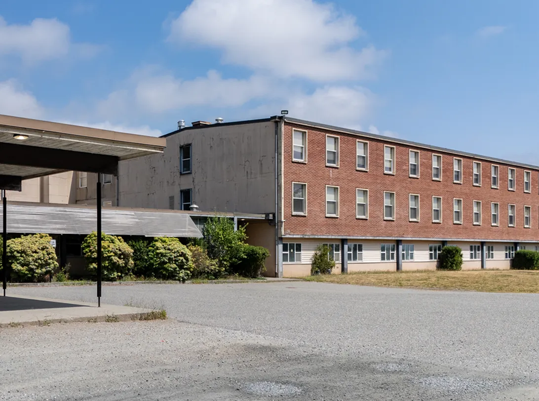 Three-story red brick and beige building with many windows under a partly cloudy blue sky, next to a gravel courtyard and some shrubs.