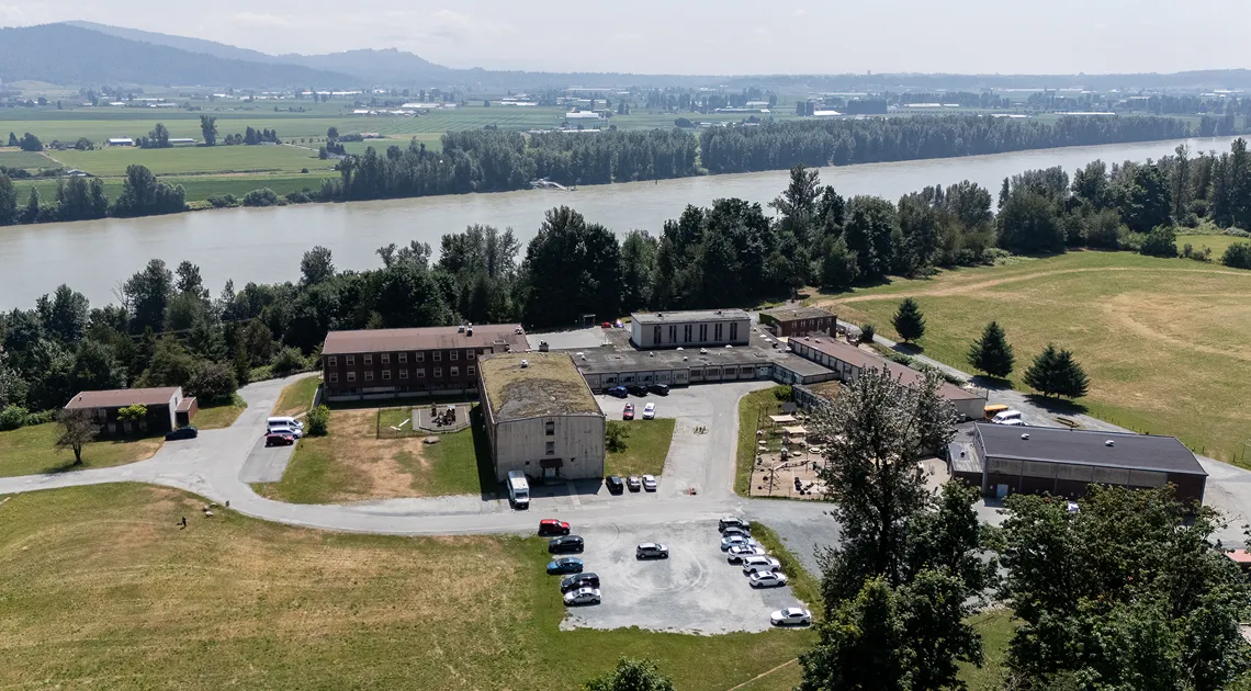 Aerial view of a rural school complex with multiple buildings, parked cars, adjacent grassy fields, and a river surrounded by trees in the background.