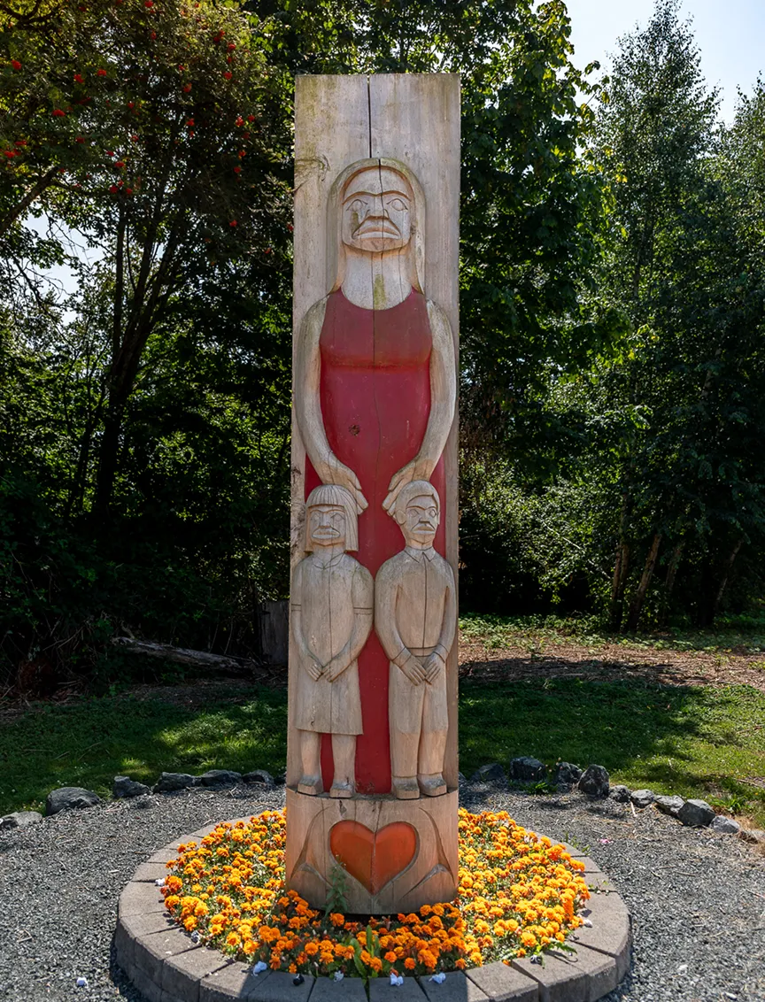 Wooden totem pole carved with figures of a tall woman in a red dress holding two smaller figures, surrounded by orange marigold flowers.
