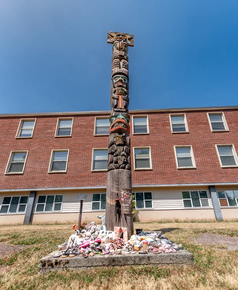 Tall carved totem pole with colorful designs standing in front of a brick building under a clear blue sky, surrounded by a memorial of small objects and flowers at its base.