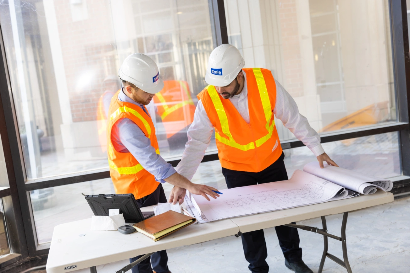 Two construction workers wearing white helmets and orange safety vests reviewing blueprints on a table inside a building under construction.