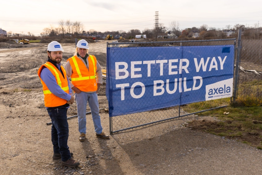 Two construction workers in orange safety vests and white helmets standing on a construction site next to a blue sign that reads 'BETTER WAY TO BUILD' with the Axela Construction logo.