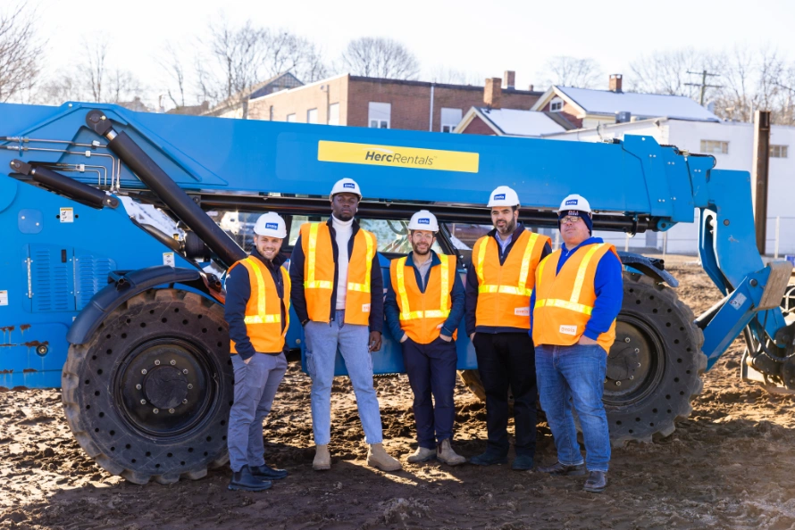 Five construction workers wearing white hard hats and orange safety vests standing in front of a large blue Herc Rentals lift on a dirt site.
