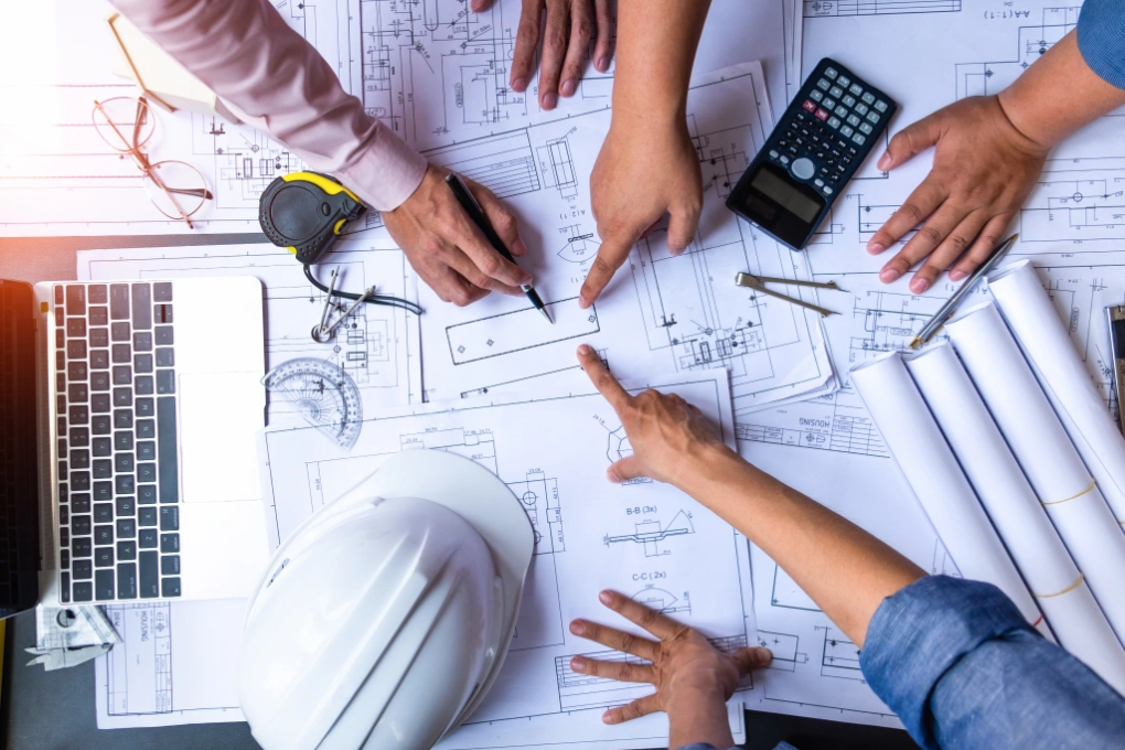 Hands of three people pointing and discussing over architectural blueprints on a table with a laptop, calculator, measuring tape, and white hard hat.
