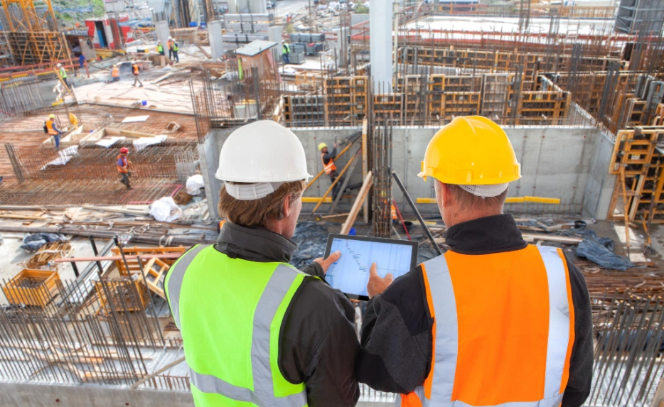 Two construction workers wearing safety vests and helmets reviewing data on a tablet at a building site.