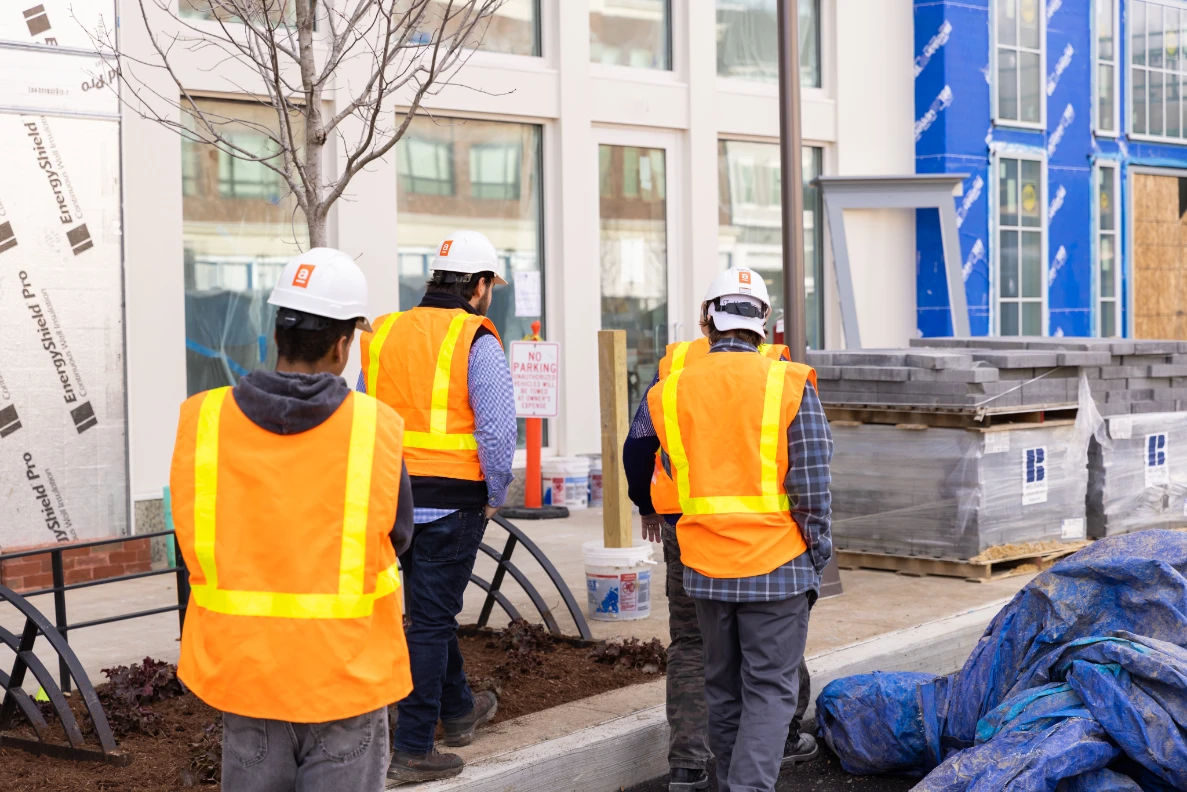 Three construction workers wearing white helmets and orange safety vests walking by a building under construction.