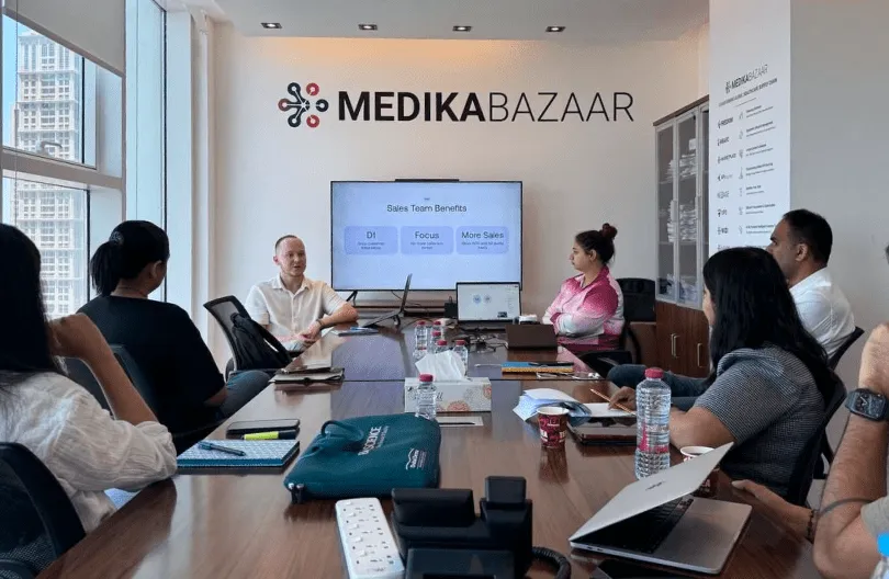 Team members sitting around a conference table in a modern office with a MEDIKABAZAAR logo and a presentation on a screen about sales team benefits.
