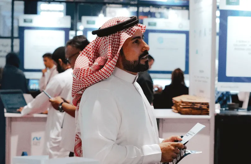 Man in traditional Middle Eastern attire holding a brochure in a busy exhibition or conference setting.