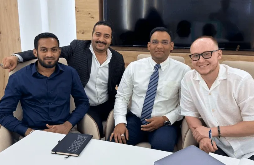 Four men sitting together in an office setting, smiling at the camera, with a notebook and laptop on the table in front of them.