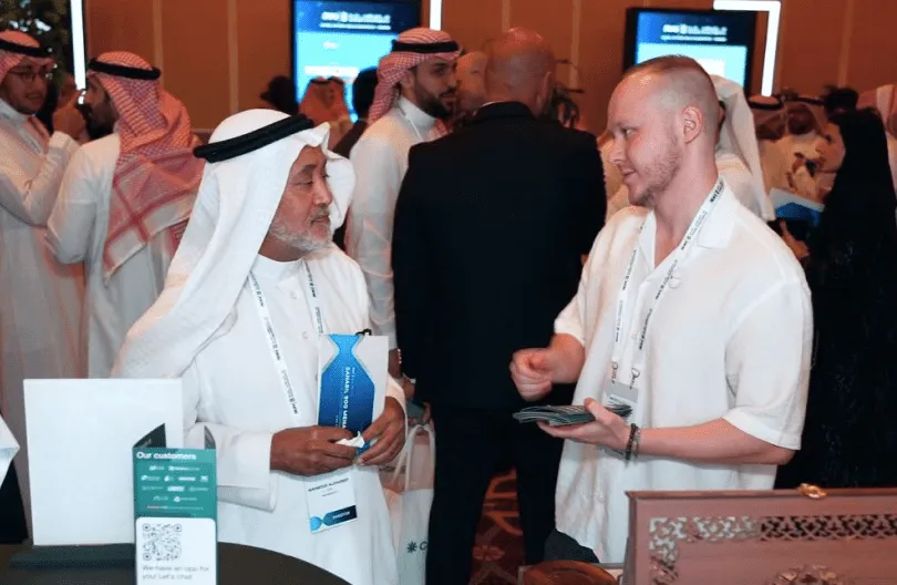 Two men engaged in conversation at an event, one wearing traditional Middle Eastern attire and the other in a white shirt, with a group of people in the background.