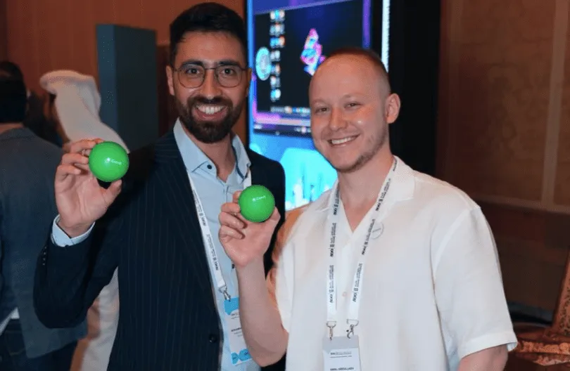 Two men smiling and holding up green stress balls at a conference or event.