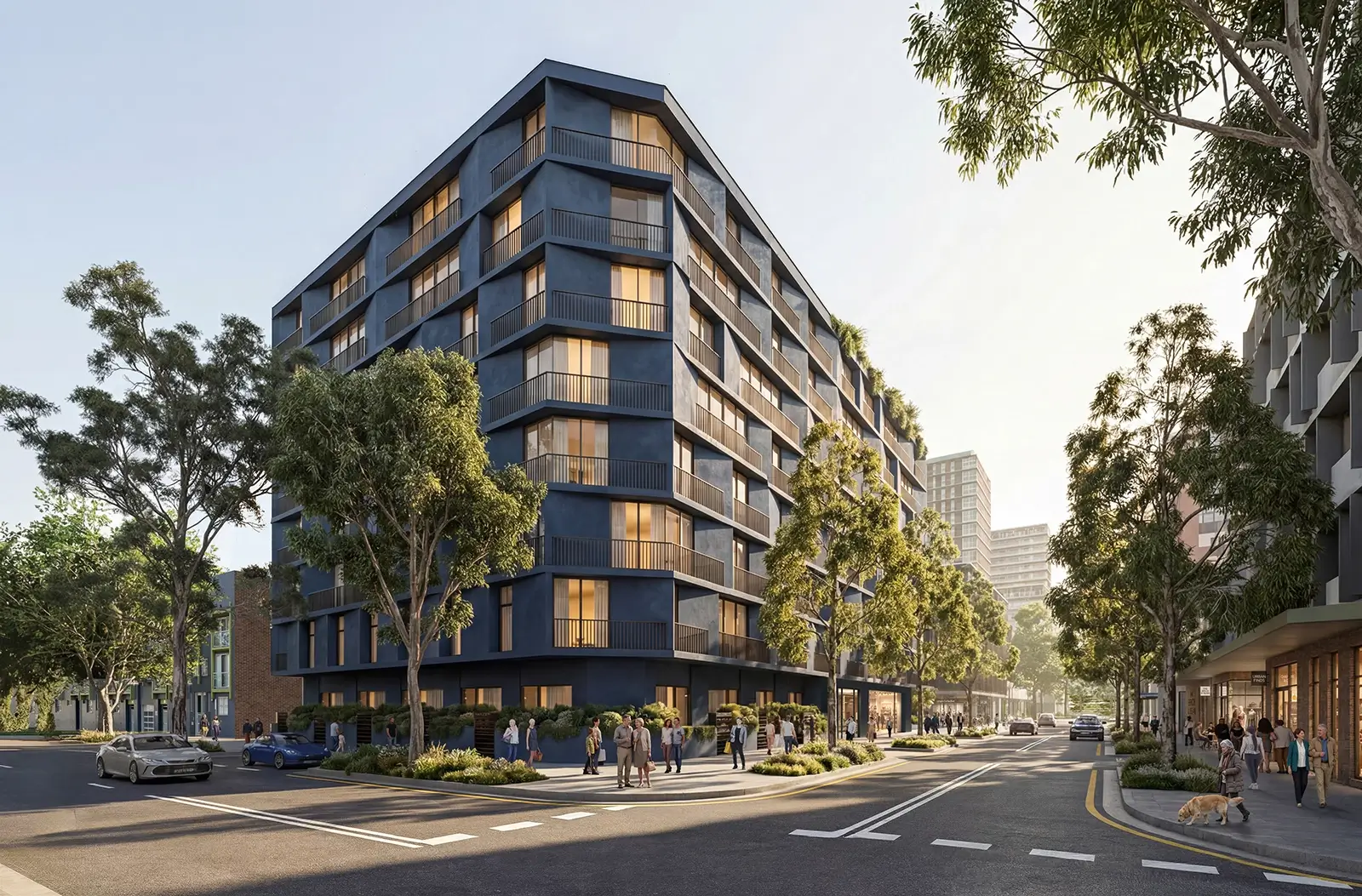 The image shows a modern, mid-rise apartment building with dark blue exterior and large windows on a street corner lined with trees. People are walking on the sidewalks, and a few cars are parked or driving on the street. The scene is bright and sunny, with additional buildings visible in the background, creating an urban neighbourhood atmosphere.