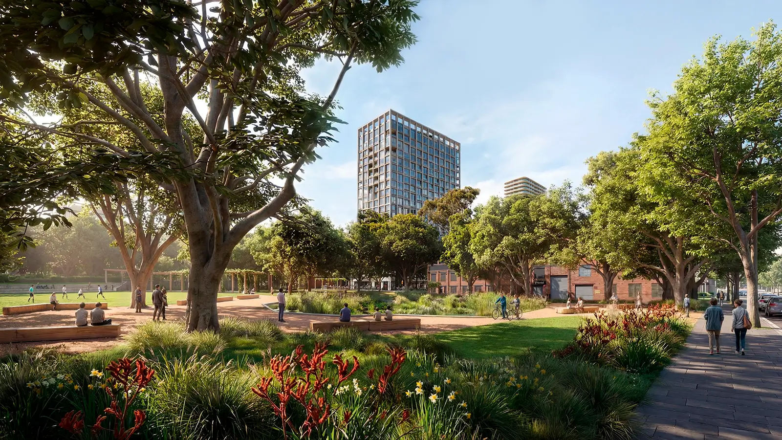 The image shows a lively park with green grass, colourful flowers, and big trees. In the front, there are red and green plants next to a paved path, where two people are walking. Around the park, people are sitting on benches, walking, cycling, and a group of children are playing on a sports field to the left. Tall trees surround the park, and in the background, there is a tall modern building with a grid pattern, along with other shorter buildings. The sky is clear and blue, and sunlight shines through the trees, making the park feel warm and welcoming. The scene shows a friendly, relaxed community in a green urban space.