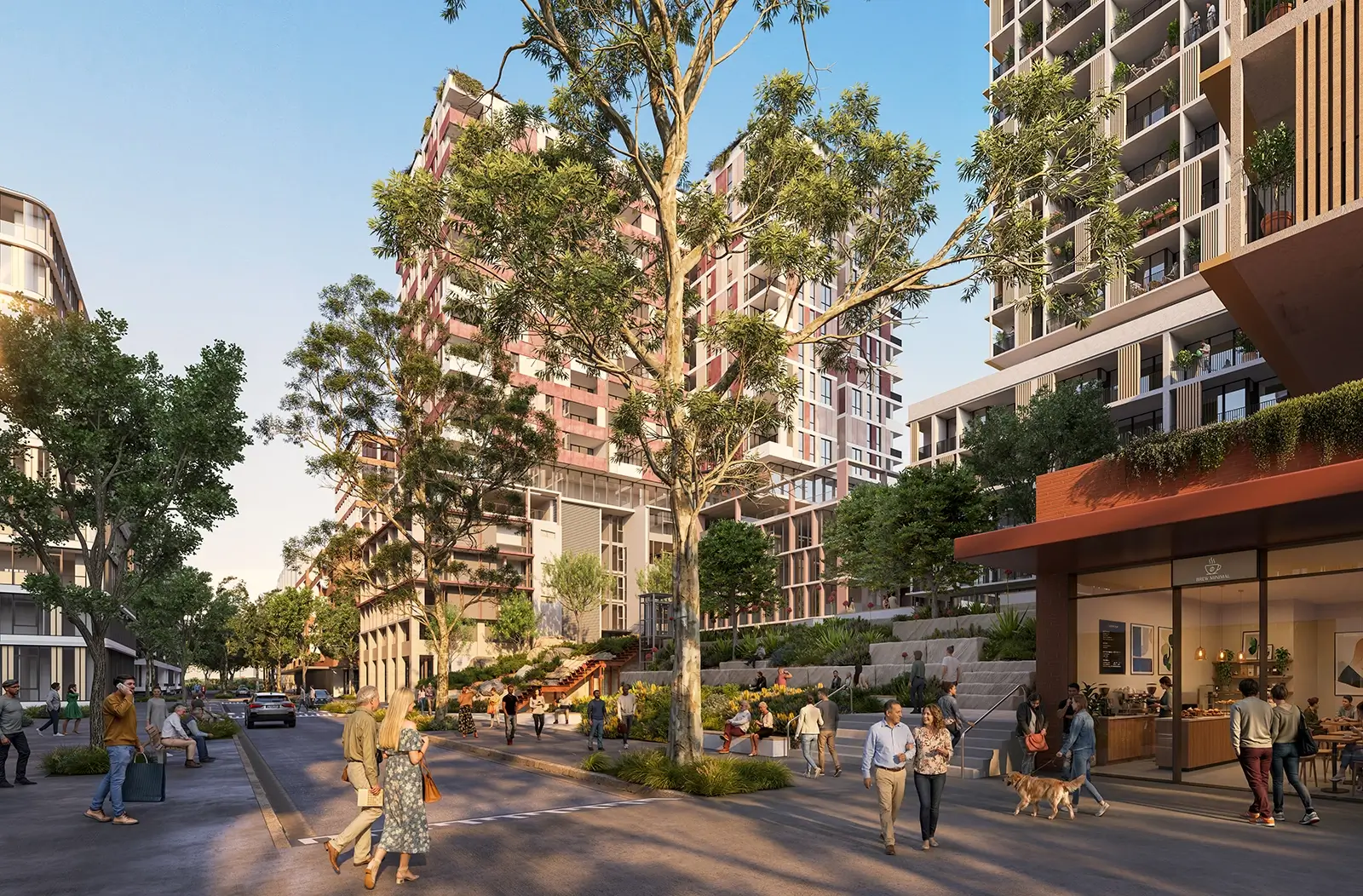 The image shows a busy, modern street during the day. There are tall apartment buildings on both sides, with big windows and balconies. Some buildings have plants on the walls and terraces. The street is wide and has many people walking, sitting on benches, and riding bikes. Some people are with friends or walking dogs. On the right, there is a café with people inside and outside, eating and drinking. Trees and garden beds line the street, adding lots of green. Steps and ramps lead up to the buildings. The place feels friendly and lively, with lots of people and plants.