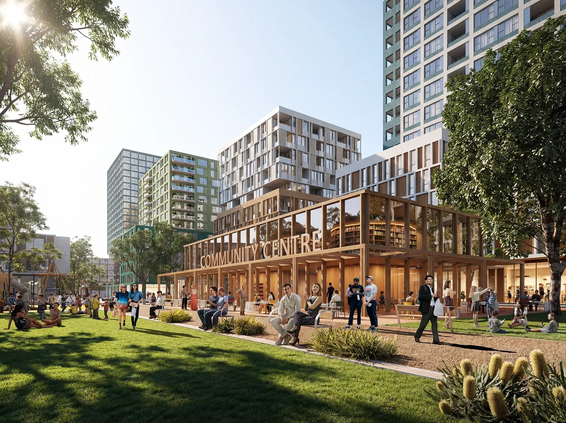 The image shows a vibrant urban park scene with people relaxing, walking, and socialising on a grassy area in front of a modern, glass-walled community centre. Surrounding the park are several tall, contemporary apartment buildings with a mix of green and white facades. Trees and landscaped plants add greenery, and the atmosphere is bright and welcoming, suggesting a lively community space.