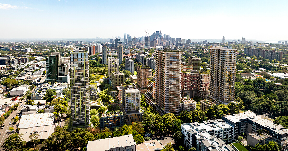 A view from above of a modern urban precinct featuring multiple high-rise residential and commercial buildings between landscaped green spaces. The area is densely built, with a mix of tall towers and mid-rise structures. In the background, a city skyline with additional skyscrapers is visible, suggesting it is close to a central business district. The overall scene conveys a vibrant, newly developed urban environment with significant integration of greenery among the buildings.