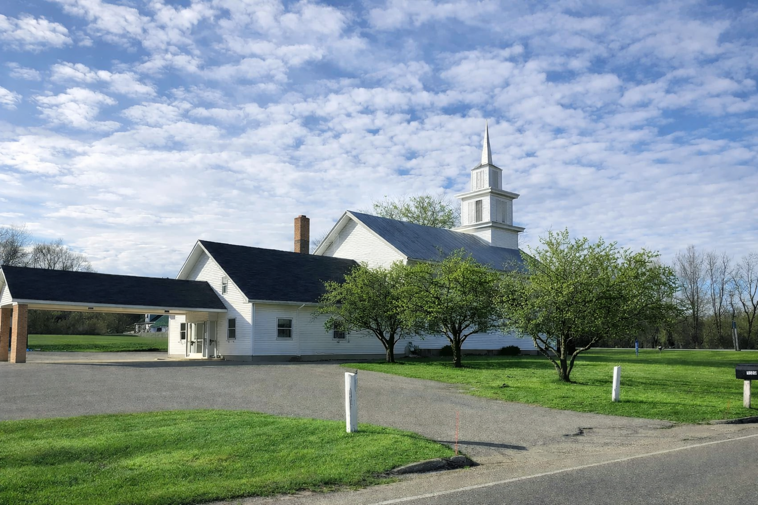 White church with a steeple surrounded by green grass and trees under a partly cloudy blue sky.