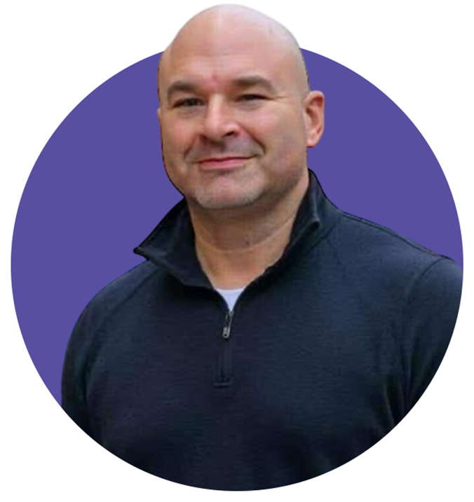 Bald man with light stubble wearing a dark zip-up sweater smiling slightly against a purple circular background.