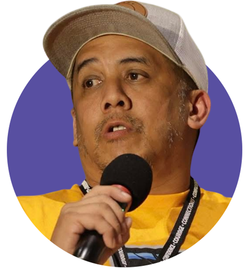 Man wearing a beige cap and yellow shirt speaking into a microphone with a dark purple background.