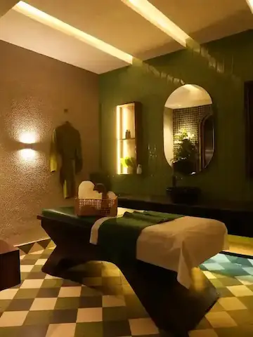 Cozy spa room with massage table covered by a white sheet and green blanket, green wall tiles, oval mirror, and soft warm lighting.