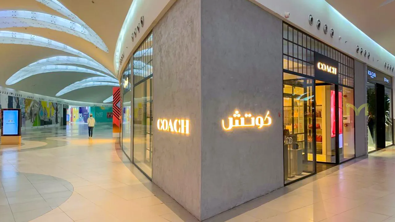 Corner of a shopping mall with illuminated Coach store signs in English and Arabic and a person walking away.