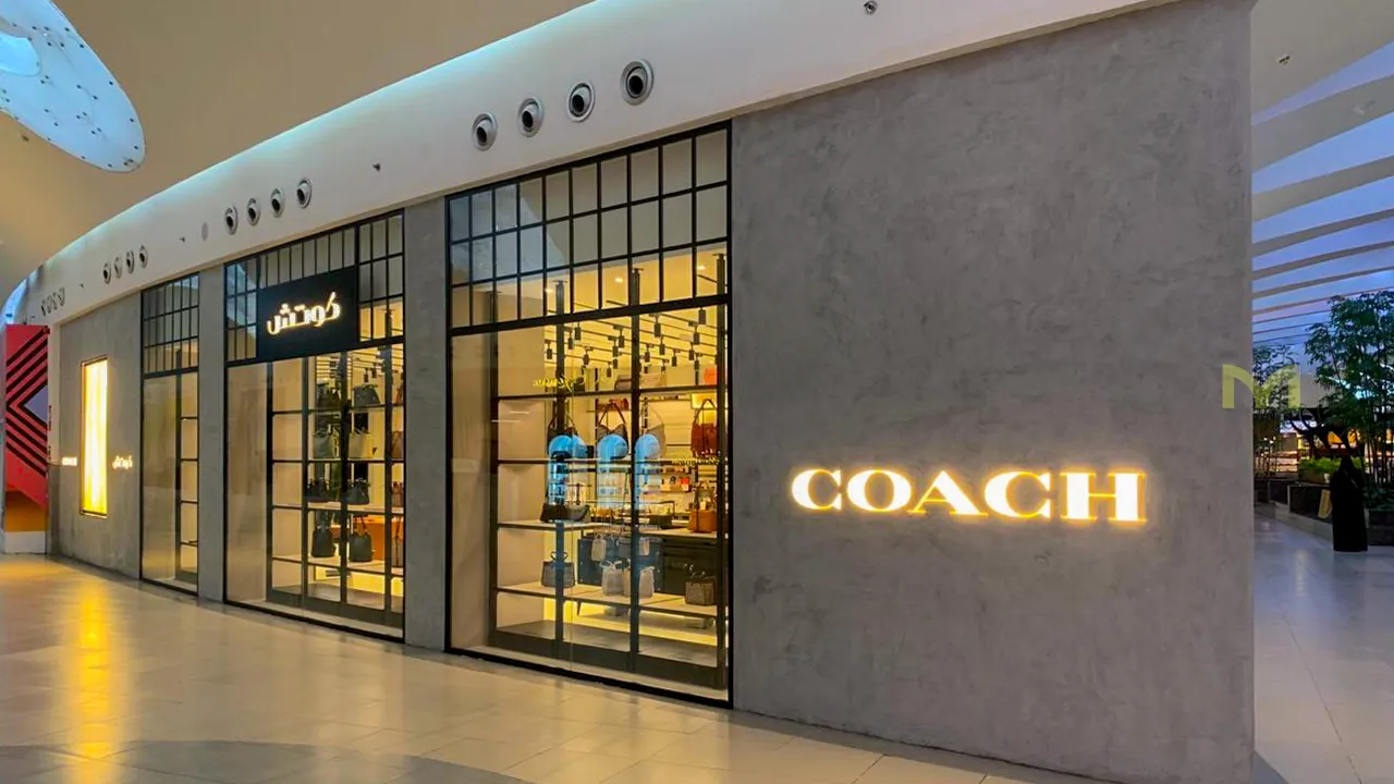 Exterior of a Coach retail store with illuminated brand sign and handbags displayed inside.